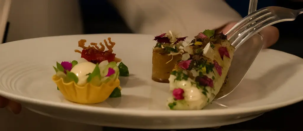 Michelin yıldızlı restoranda sunulan dört parçalık fine dining tadım tabağı; çiçek yapraklarıyla süslenmiş balık, mini tart, domatesli krokan ve silindirik amuse-bouche