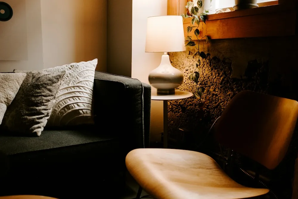 Cocooning etkisi taşıyan salon köşesi: sıcak masa lambası aydınlatması, dokulu yastıklar, doğal ahşap yüzey ve bitki detaylarıyla samimi ve huzurlu ev atmosferi.