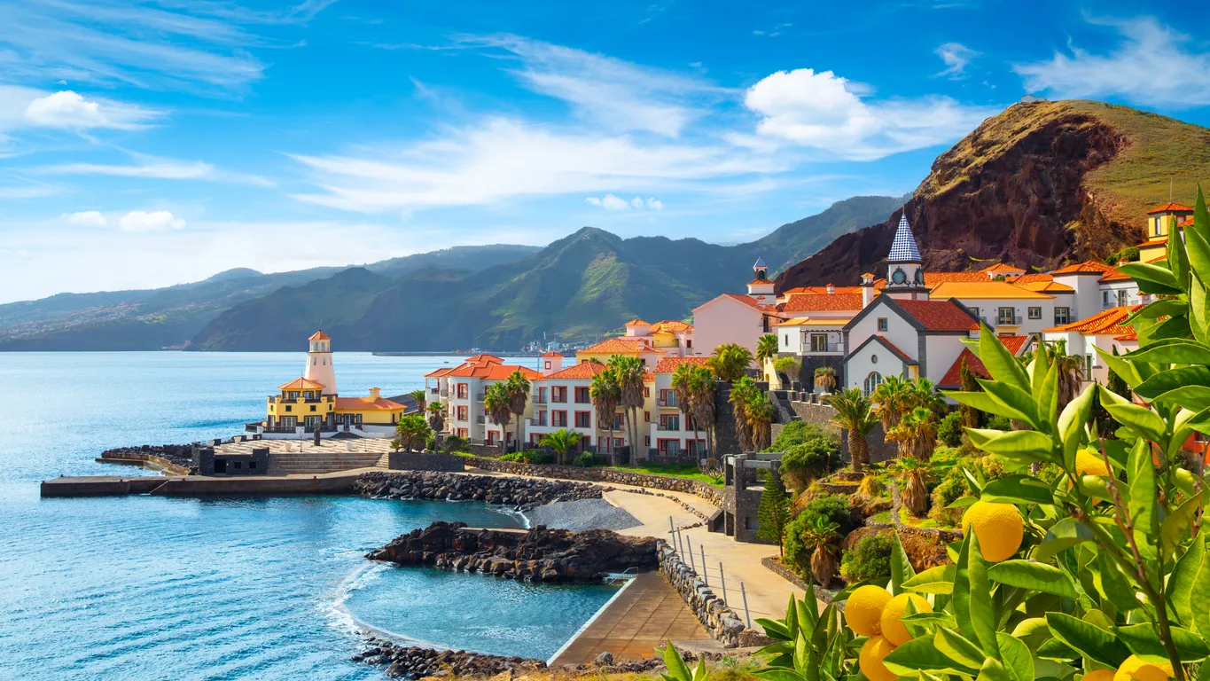 Mavi gökyüzü ve turkuaz sularla çevrili Madeira sahilinde, yemyeşil dağların eteğinde, turuncu kiremitli çatıları ve palmiye ağaçlarıyla bezeli şirin evlerin bulunduğu, sol tarafta bir deniz feneri ve sağda meyve yüklü bir ağacın kadraja girdiği pitoresk bir kasaba manzarası.