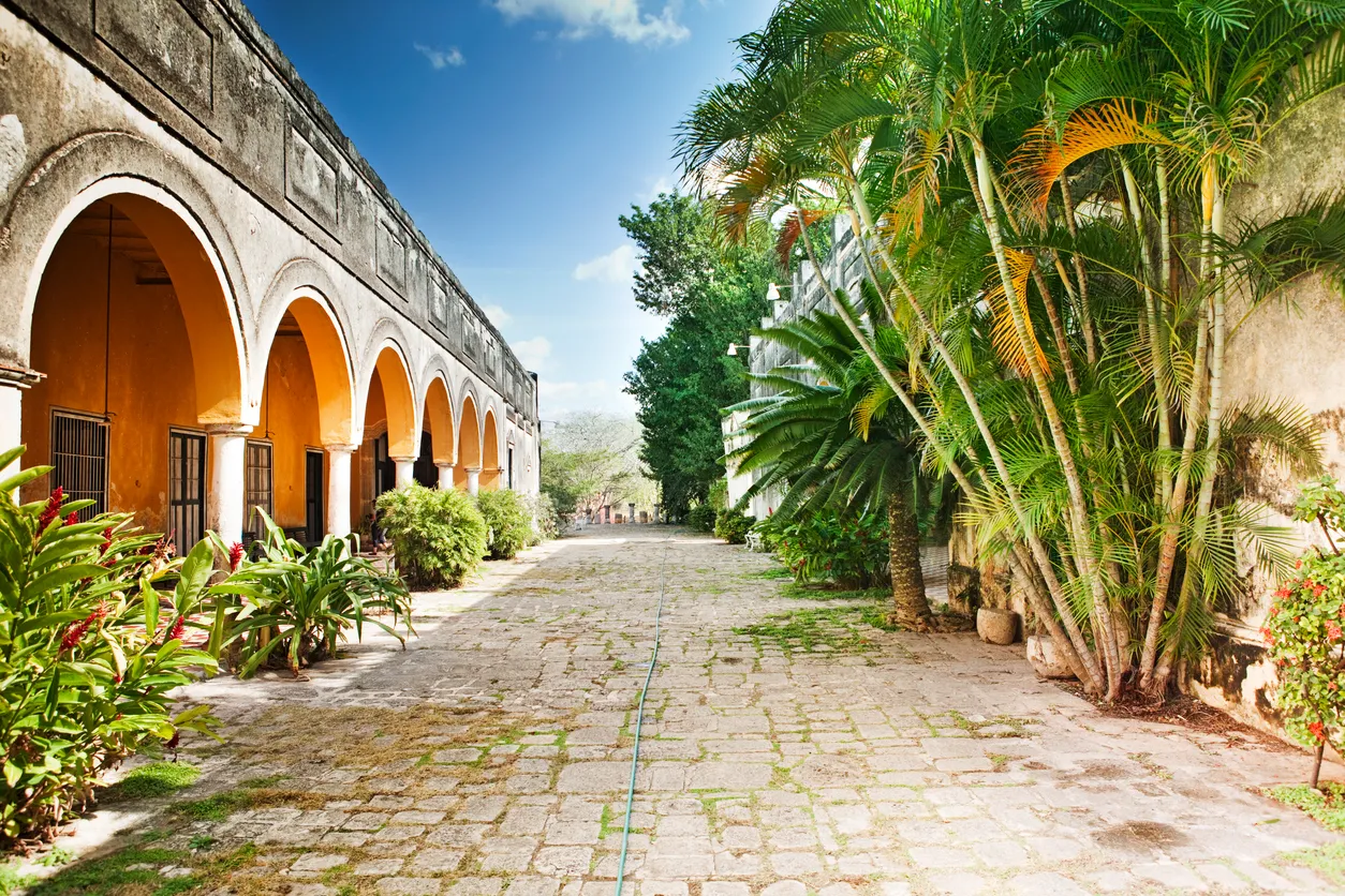 Kemerli, sarı cepheli tarihi bir binanın solunda, taş döşeli bir yolun sağında ise yemyeşil palmiye ağaçları ve tropik bitki örtüsünün sıralandığı bu huzurlu Yucatan manzarası, masmavi gökyüzünün altında uzanıyor ve bölgenin eşsiz atmosferini hissettiriyor.
