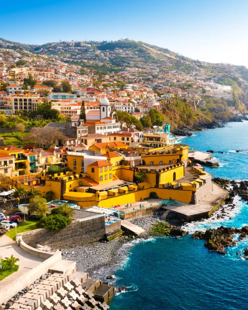 Masif sarı bir kale yapısının etrafında, yamaç boyunca sıralanmış kırmızı çatılı evleri ve beyaz kulesiyle masmavi denizin kıyısında uzanan, huzurlu tatil rotaları için ideal, yemyeşil tepelere kurulu canlı bir sahil kasabası panoraması.