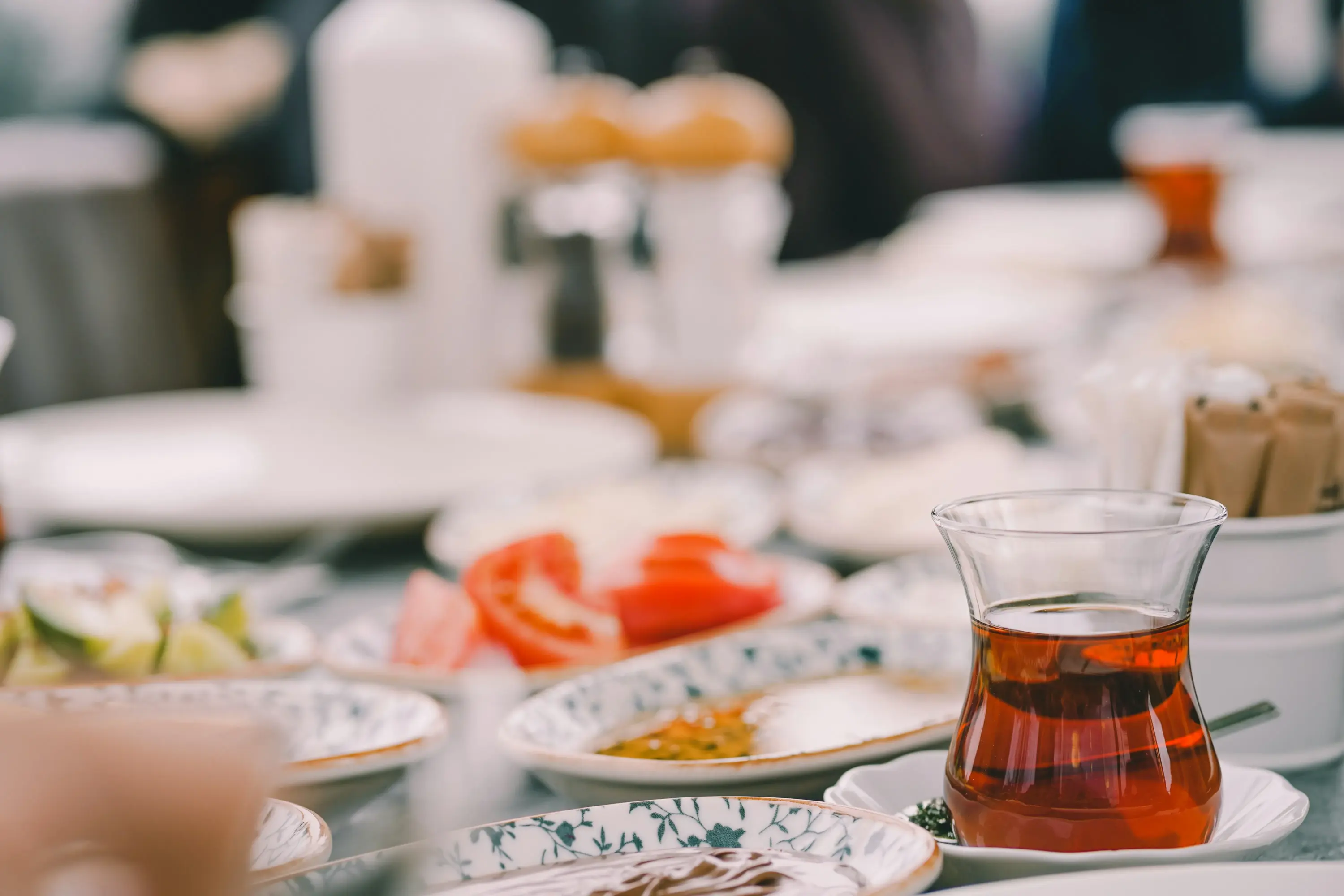 Yakın çekimde, geleneksel ince belli bir çay bardağında taze demlenmiş çay, dilimlenmiş domates, salatalık, çikolata kreması ve çeşitli yöresel lezzetlerin bulunduğu, Şef Sezai Erdoğan'ın restoran önerileri bağlamında keyifli bir kahvaltı sofrası görünüyor.
