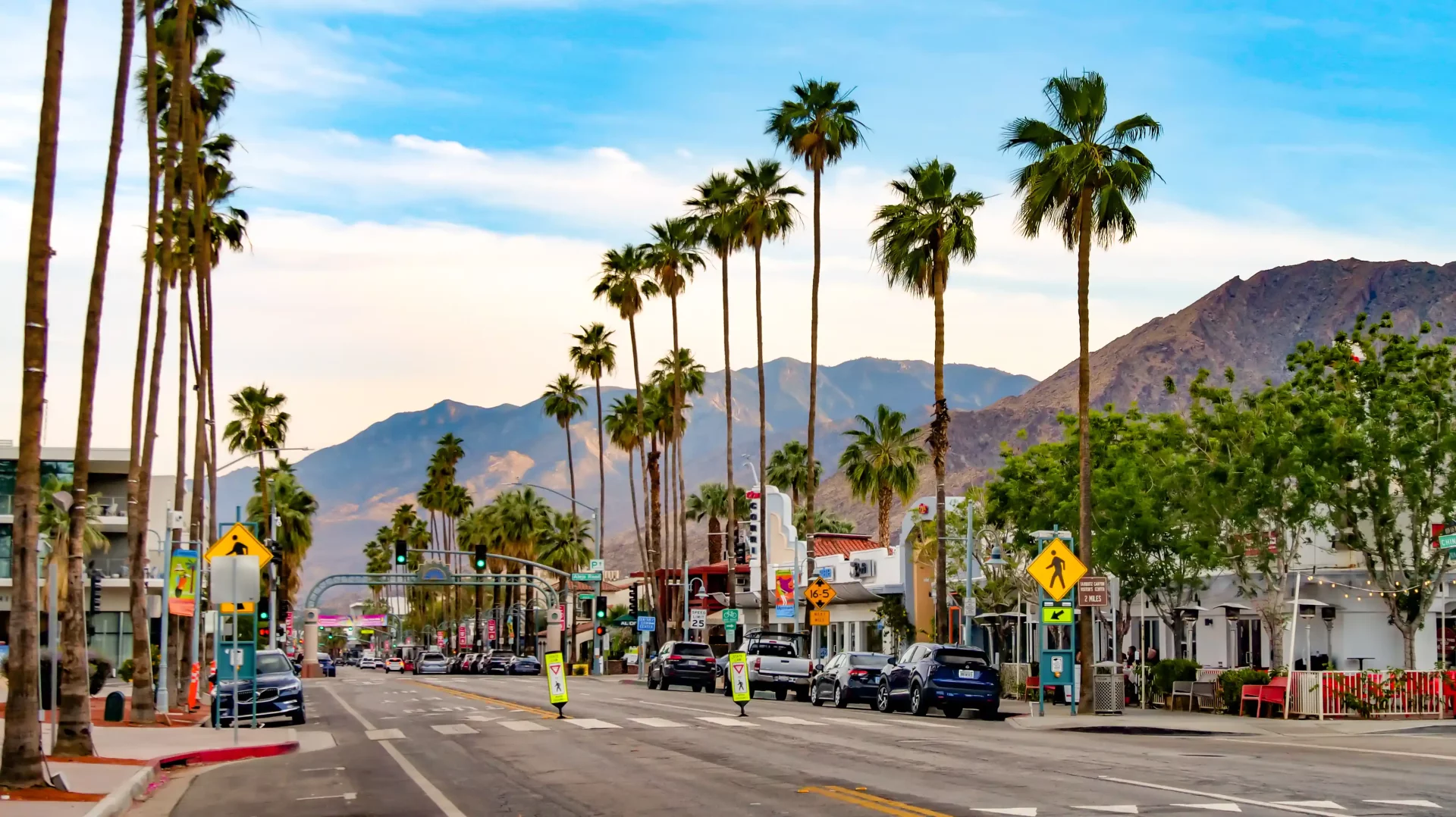 Canlı bir atmosfer sunan Palm Springs'teki geniş bir caddenin güneşli görüntüsünde, kaldırım kenarlarında sıralanan uzun palmiye ağaçları, yola park etmiş ve hareket halinde olan arabalar ile dükkanlar ve arka plandaki dağlar net bir şekilde görülüyor.