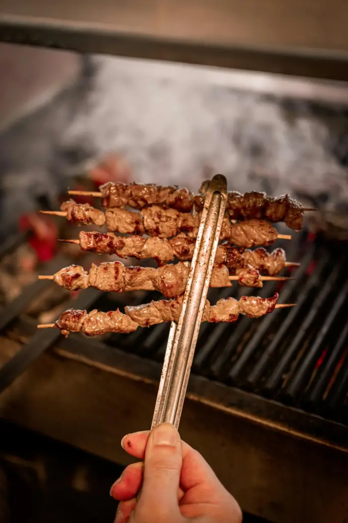Köz ateşinde ızgarada pişirilen, metal maşayla tutulan lezzetli dana et şiş kebapları yakın çekim, cağ kebabı.