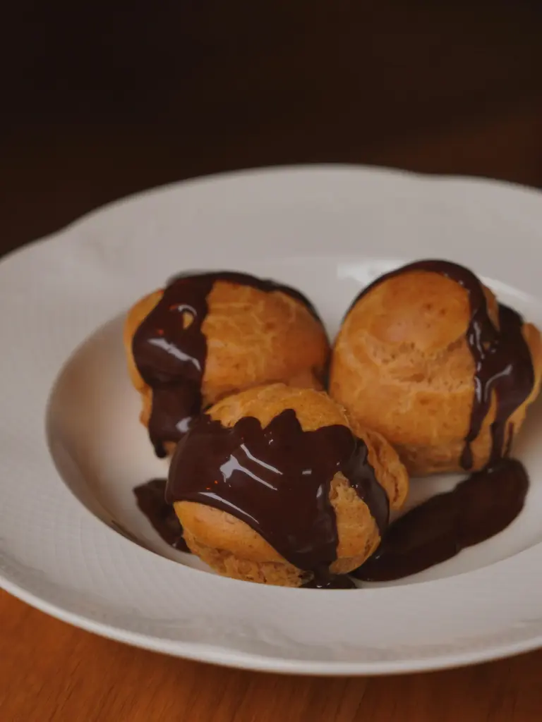 Beyaz tabakta üç adet profiterol üzerine dökülmüş bitter çikolata sosu ile servis edilmiş.
