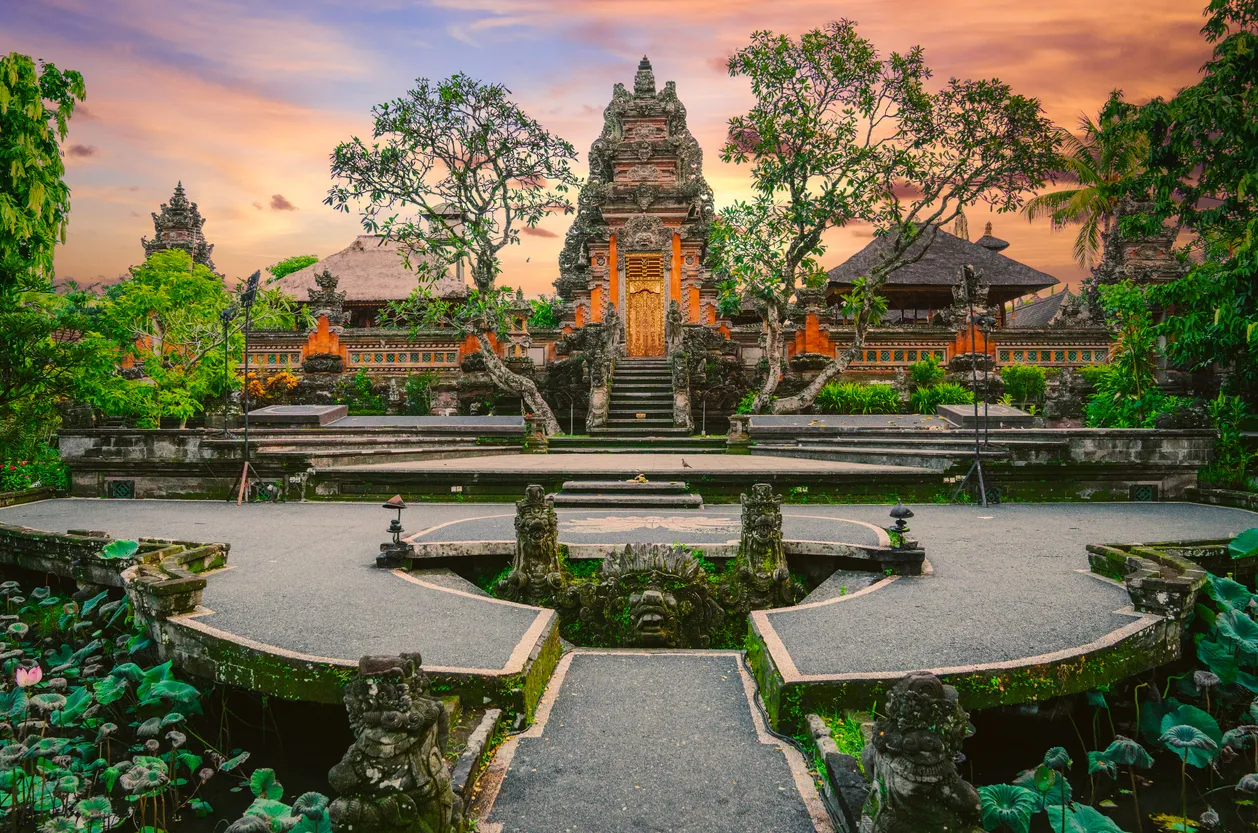 Ubud'daki geleneksel Bali tapınağının görkemli, oymalı ana kapısı, yeşil ağaçlarla çevrili ve altın renkli bir kapıya sahip; ön planda nilüfer yaprakları ve birkaç pembe nilüfer çiçeğiyle dolu bir gölet ile süslü taş heykellerin ve bir devasa yüz oyma detayıyla çevrili, portakal rengi gün batımı gökyüzünün altında çarpıcı bir şekilde yükseliyor.