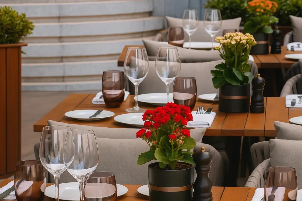 St. Regis Brasserie ilhamlı şık bir açık hava restoranı; ahşap masalarda beyaz tabaklar, şarap kadehleri, renkli bardaklar ve çiçek aranjmanlarıyla zarif bir şekilde düzenlenmiş oturma alanı.