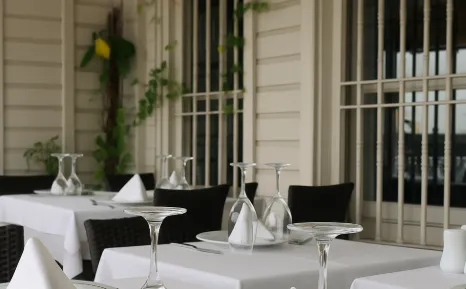 Arnavutköy’deki Sur Balık restoranının beyaz masa örtülü şık dış mekân oturma alanında ilhamlı mekan; zarif tabak sunumları, ters çevrilmiş şarap kadehleri ve yeşil sarmaşıklarla çevrili ferforjeli pencere önünde huzurlu bir atmosfer.