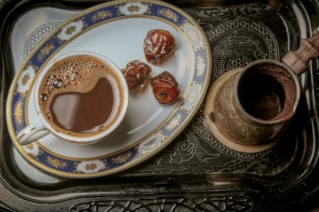 Gümüş tepsi üzerinde geleneksel fincanda sunulan köpüklü Türk kahvesi, yanında hurmalar ve işlemeli cezve ile zarif bir sunum.