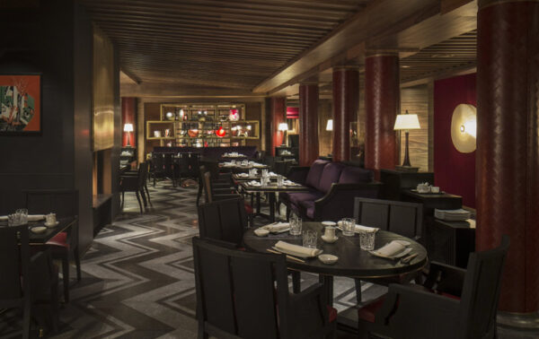Four Seasons Megéve kayak otelinin içindeki lüks restoran, koyu kırmızı sütunlar, zikzak desenli zemini, ahşap tavanı ve mor banket koltuklarıyla donatılmış masalarıyla zarif bir atmosfer sunuyor.
