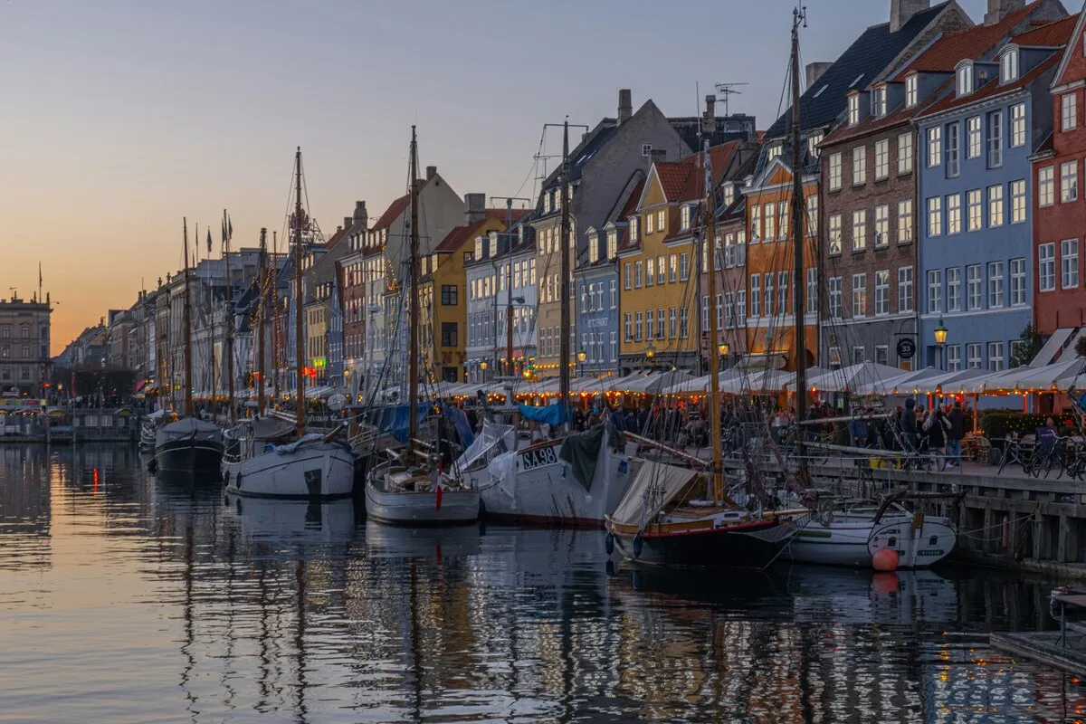 Kopenhag'ın ikonik Nyhavn kanalında, alacakaranlığın pastel tonları altında, çok sayıda yelkenli tekne suya yansırken, kıyı şeridindeki canlı renkli tarihi binaların pencereleri ve kafe tentelerinin altındaki ışıklar keyifli bir akşam atmosferi sunuyor.