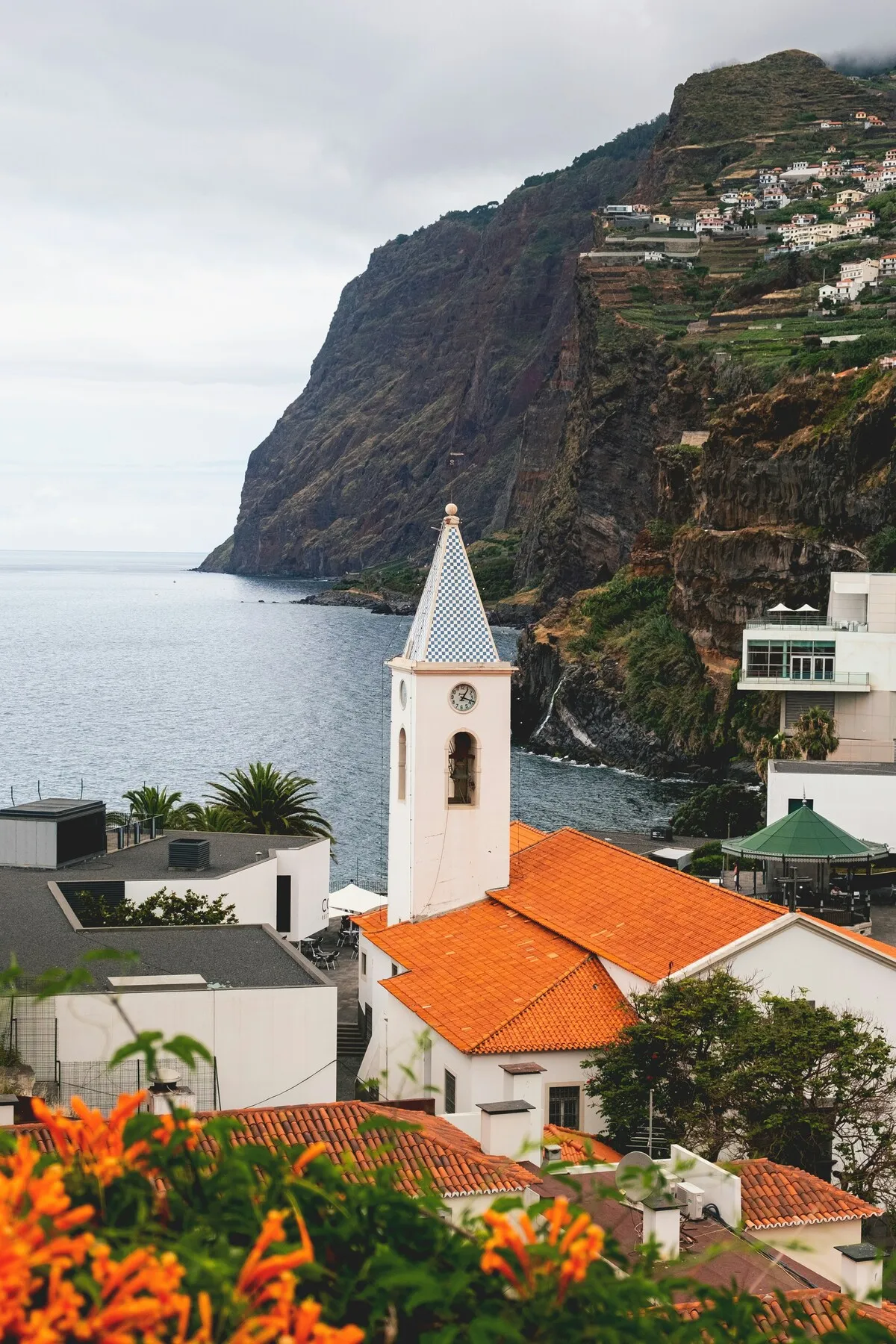 Madeira, Portekiz'in büyüleyici kıyı şeridini yansıtan bu fotoğrafta, mavi-beyaz damalı sivri çatılı beyaz bir kilise kulesi ön planda dikkat çekerken, arkasında uzanan dingin masmavi okyanus, dramatik sarp kayalıklar ve yamaçlara serpiştirilmiş geleneksel evler, ön plandaki turuncu çiçeklerle pitoresk bir manzara oluşturuyor.