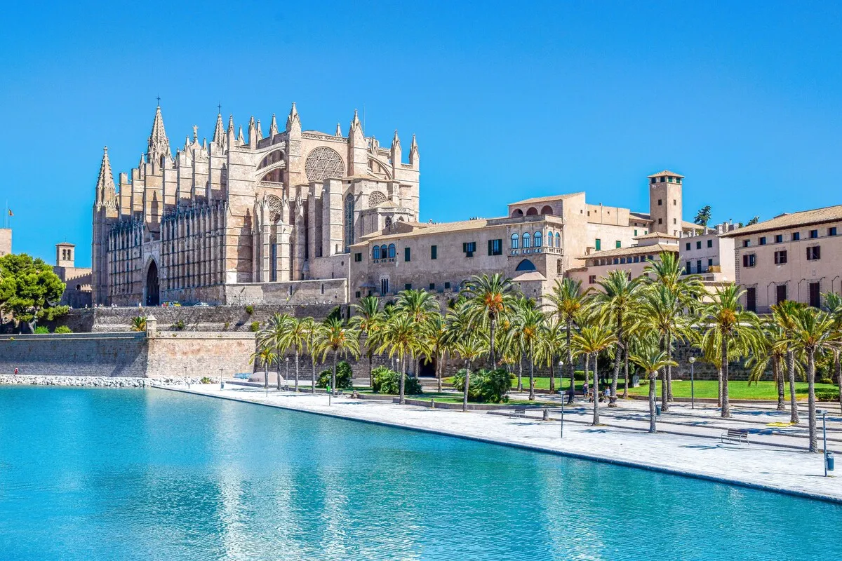 Palma de Mallorca'daki Santa Maria Katedrali'nin masmavi bir su kütlesinin kenarında yükselen görkemli gotik yapısı, sıralanmış palmiye ağaçları ve çevredeki tarihi binalarla güneşli bir günde muhteşem bir şehir manzarasını gözler önüne seriyor.
