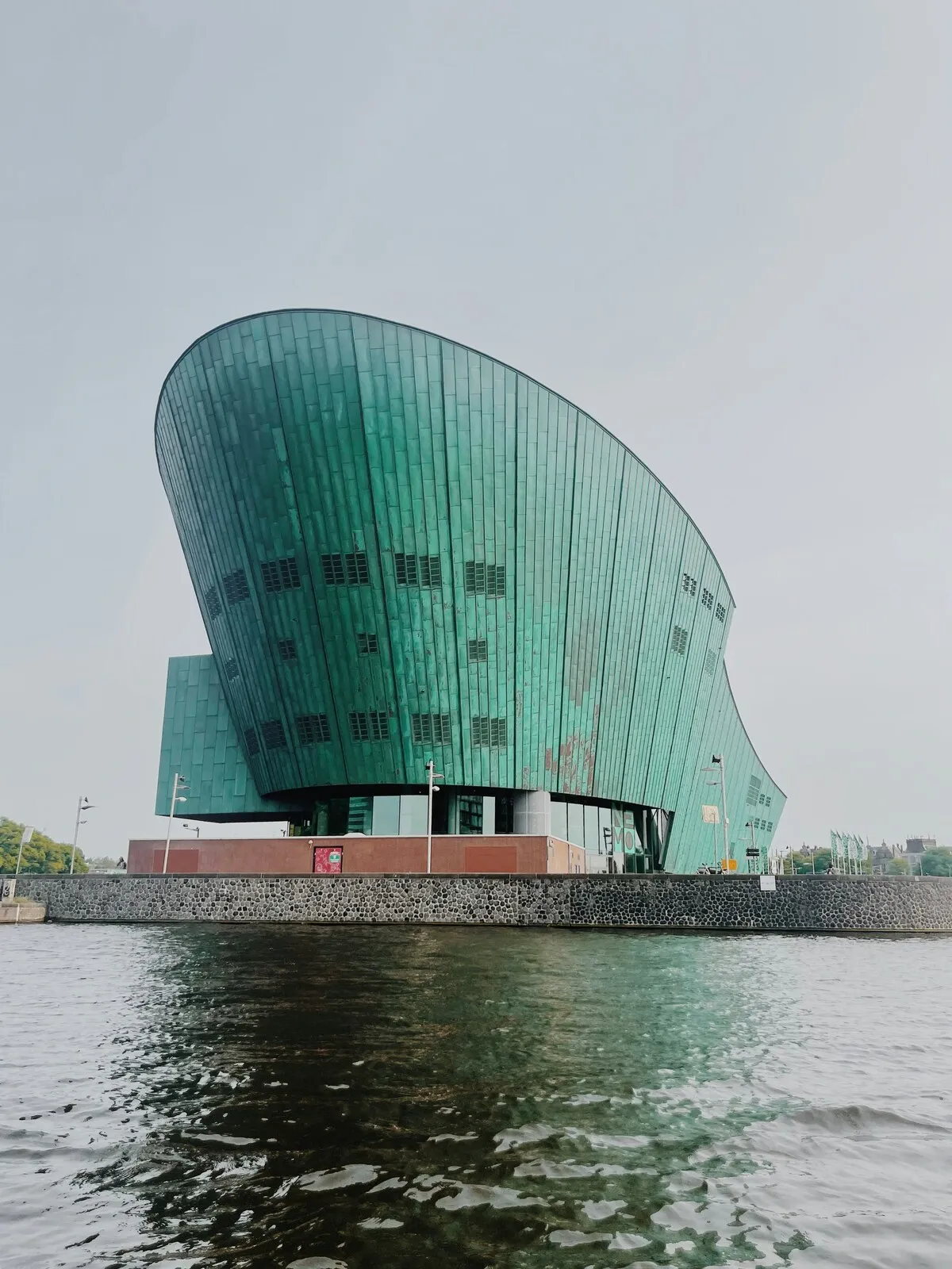 Amsterdam'da suyun üzerinde yükselen, çağdaş mimarisi ve yeşile dönmüş bakır kaplamasıyla dikkat çeken, gemiyi andıran kavisli yapıya sahip Nemo Bilim Müzesi, gri gökyüzünün altında belirgin bir şekilde görünüyor.