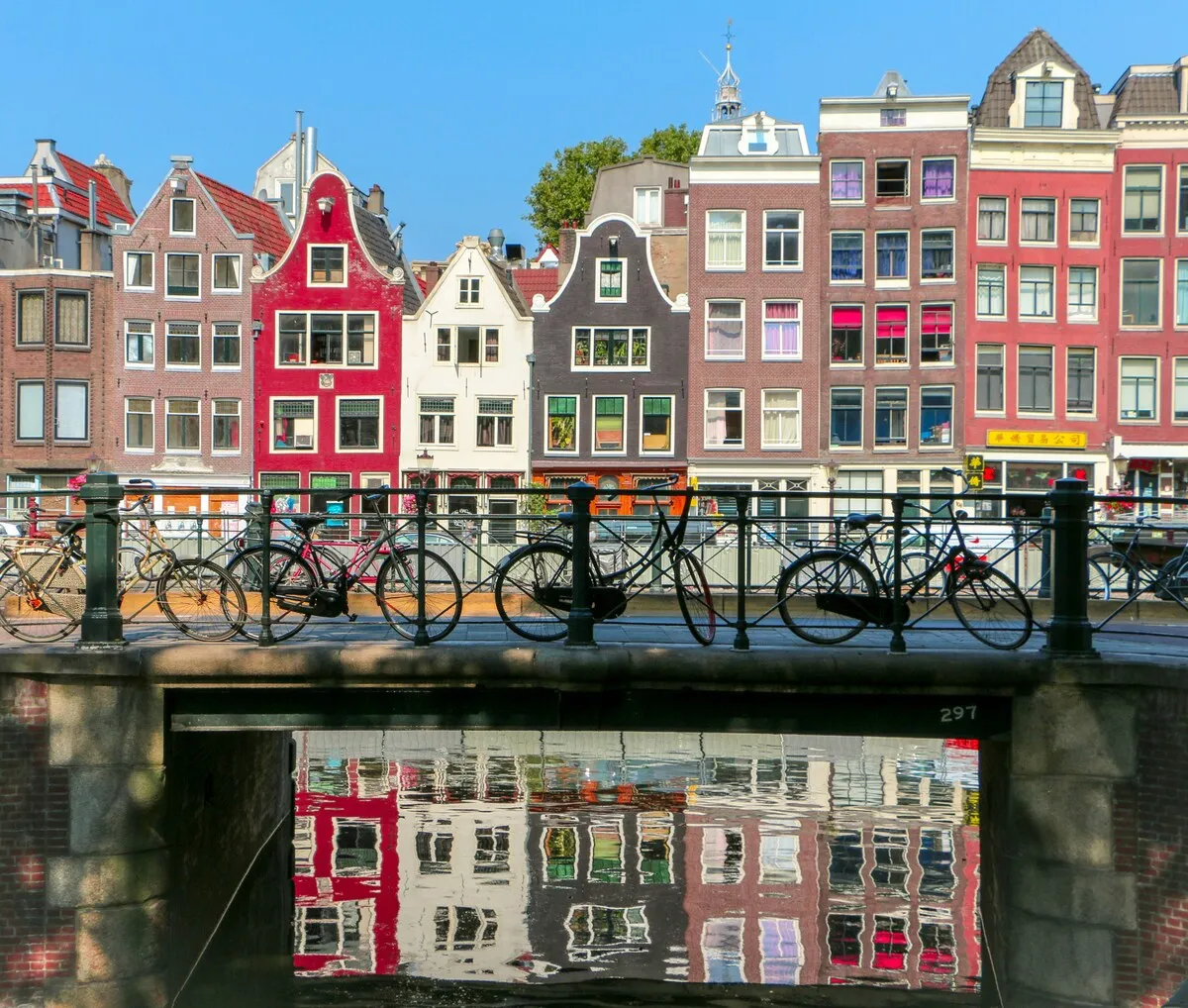 Amsterdam'ın tarihi kanal evlerinin suya yansıdığı, neşeli renkte binaların önünde sıralanmış köprünün üzerinde çok sayıda bisikletin görüldüğü güneşli bir Amsterdam tatili sahnesi.