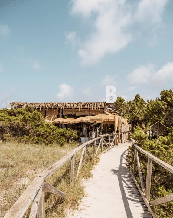 Güneşli bir günde, Formentera'daki Beso Beach restoranına giden, iki yanı ahşap korkuluklarla çevrili, kumlu ve otlu doğal bir patika, keyifli bir mavi yolculuk deneyiminin ardından lezzetli yemekler sunan bu özel mekana davetkar bir görünüm katıyor.