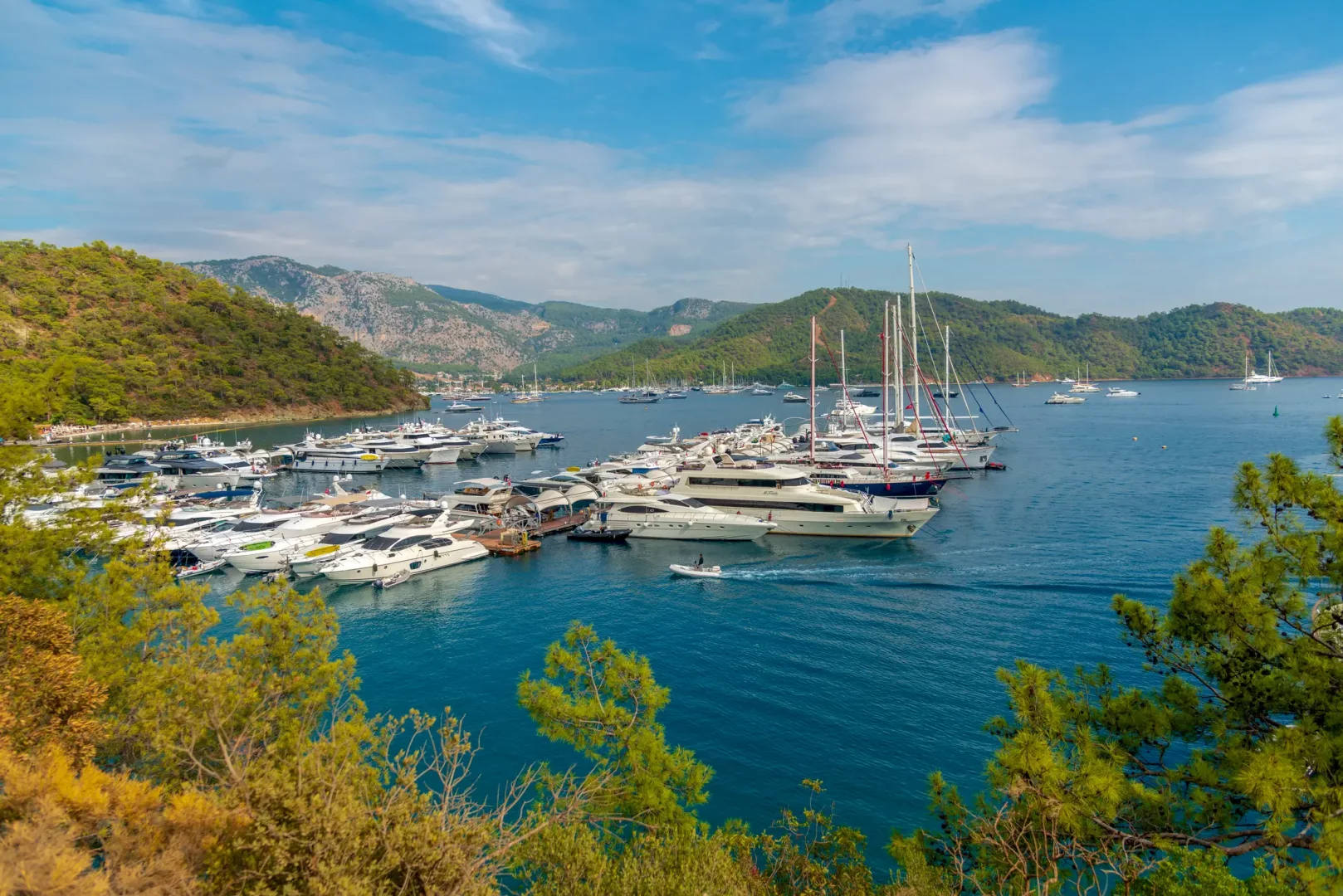 Göcek'in yemyeşil tepelerle çevrili masmavi koyunda, farklı boyutlardaki lüks süperyatlar ve tekneler berrak denizde demirlemiş vaziyette, başarılı tasarımcı Rıza Tansu'nun bahsettiği mavi yolculuk tutkusunu ve bölgenin eşsiz güzelliğini gözler önüne seriyor.