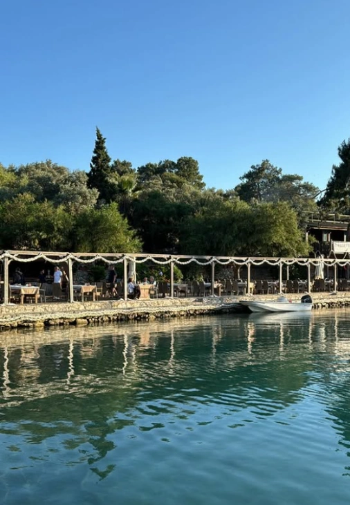 Göcek'te, Ömer Malaz'ın mavi yolculuk rotalarındaki favori durağı olan Göbün restoranının denize sıfır terasında misafirler otururken, berrak suya yansıyan yemyeşil ağaçlar ve yanaşmış bir tekneyle huzurlu bir manzara sunuluyor.