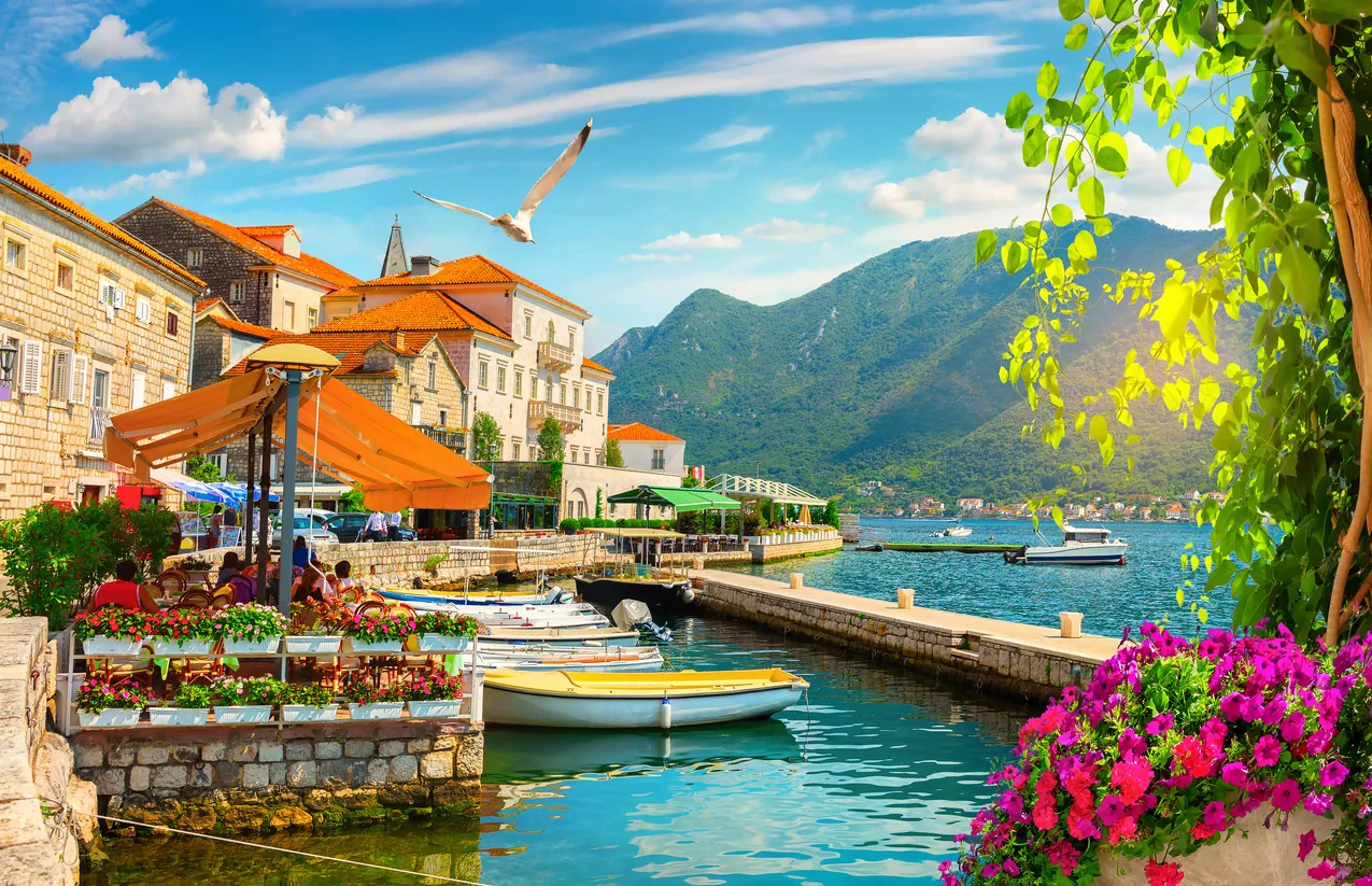 Kotor Körfezi'nin berrak sularında yansıyan, kırmızı çatılı tarihi taş binalarıyla ve etrafını saran heybetli dağlarla ünlü pitoresk Kotor şehrinin genel görünümü, güneşli bir günde büyüleyici bir doğal ve kültürel miras manzarası sunuyor.