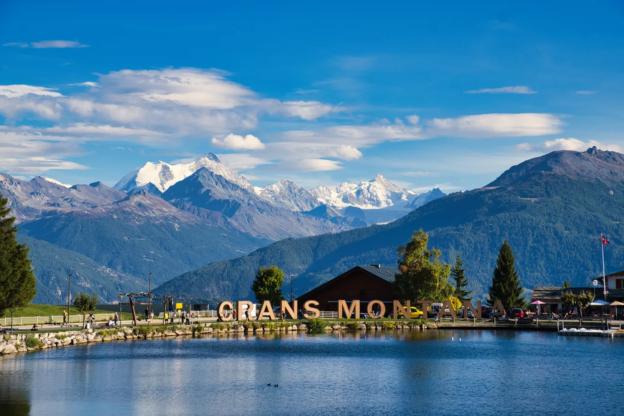 Crans-Montana’da göl kenarında yer alan yerleşim alanı, arka planda karla kaplı İsviçre Alpleri ve dağ manzarası.