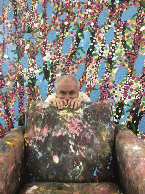 Damien Hirst, boya lekeleriyle kaplı koltukta otururken – sanatçının üretim sürecine tanıklık eden samimi bir stüdyo karesi.