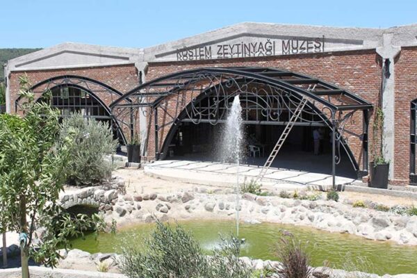 Alaçatı Rehberi'nde ziyaret edilmesi önerilen, tuğla cepheli, metal kemerli geniş girişlere sahip modern Köstem Zeytinyağı Müzesi'nin önünde, ortasında fıskiyenin yükseldiği bir havuz ve yeşil bitkilerin bulunduğu özenle düzenlenmiş bir bahçe görünüyor.