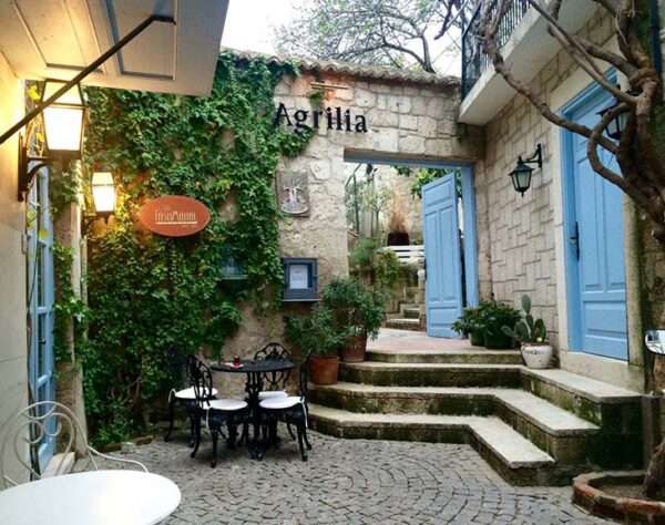 Alaçatı rehberi kapsamında önerilen Agrilia restoranının, taş duvarları yemyeşil sarmaşıklarla kaplı, açık mavi ahşap kapıları ve arnavut kaldırımlı avlusunda dört kişilik siyah ferforje masa ve sandalyelerle bezenmiş şirin dış mekanı.