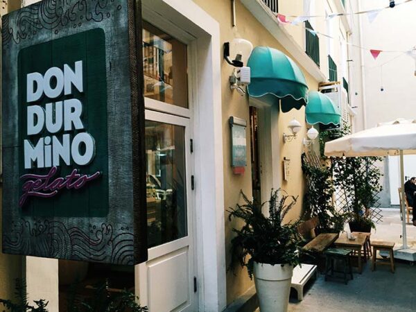 Alaçatı Rehberi'nde gelato için önerilen Dondurmino'nun dar bir sokakta konumlanmış, üzerinde büyük yeşil bir tabelası ve pastel sarısı binasıyla dikkat çeken, teal renkli tenteleri ve bitkilerle süslenmiş dış mekan oturma alanlarına sahip çekici bir görünümü.