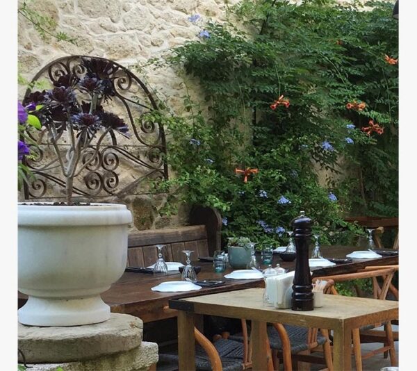 Alaçatı'da bir restoranın taş duvarlı, yemyeşil sarmaşıklar ve rengarenk çiçeklerle çevrili, üzerinde tabaklar ve kadehlerle şık bir şekilde hazırlanmış ahşap masaların bulunduğu davetkar dış mekan yemek alanı.