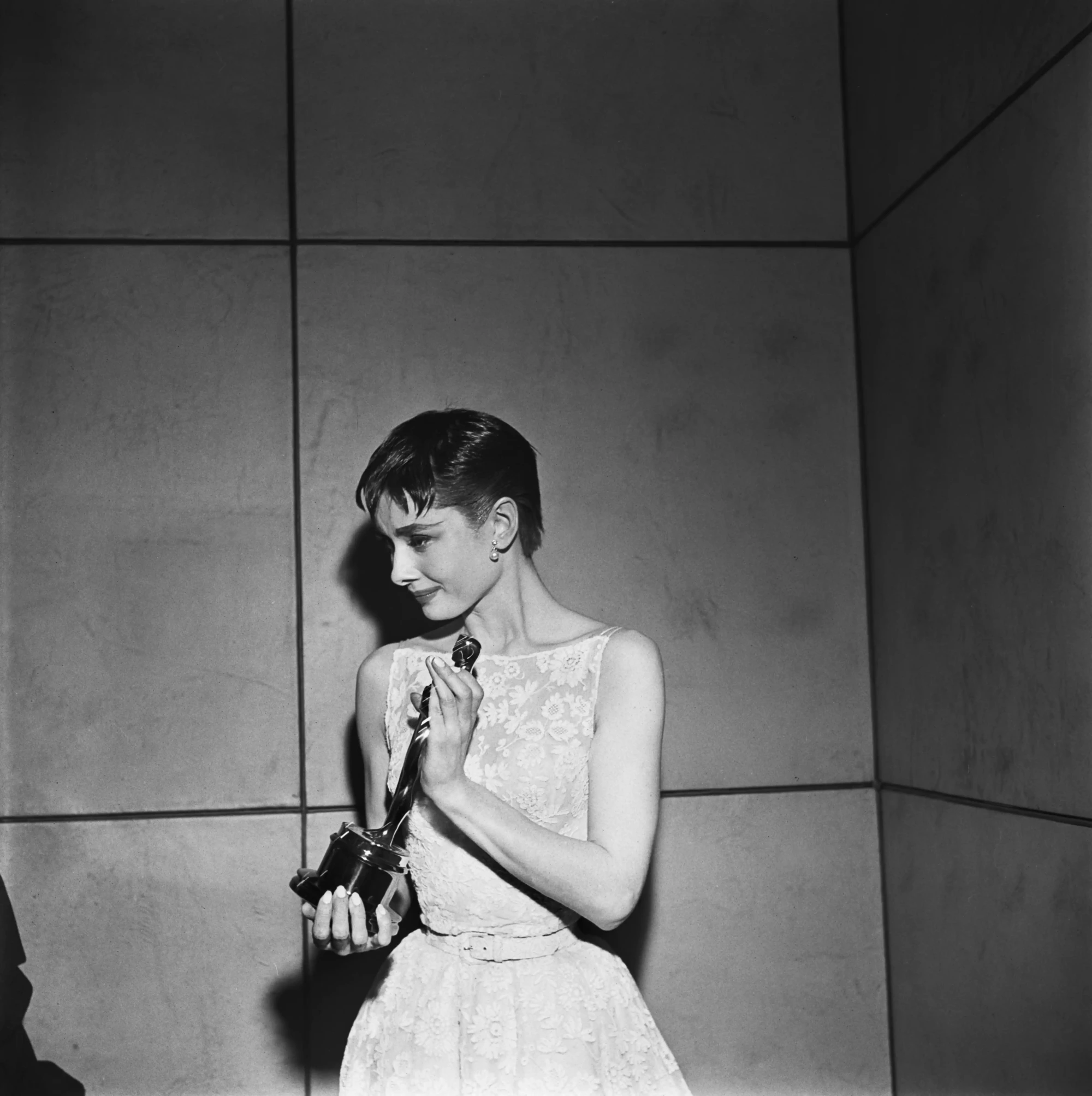 Audrey Hepburn, 1954 Oscar Ödül Töreni'nde Roman Holiday filmindeki prenses rolüyle kazandığı En İyi Kadın Oyuncu ödülünü, zarif dantelli elbisesi, kısa saçları ve inci küpeleriyle, hafifçe gülümseyerek ellerinde tutarken tarihe geçen unutulmaz anlarından birini yaşıyor.