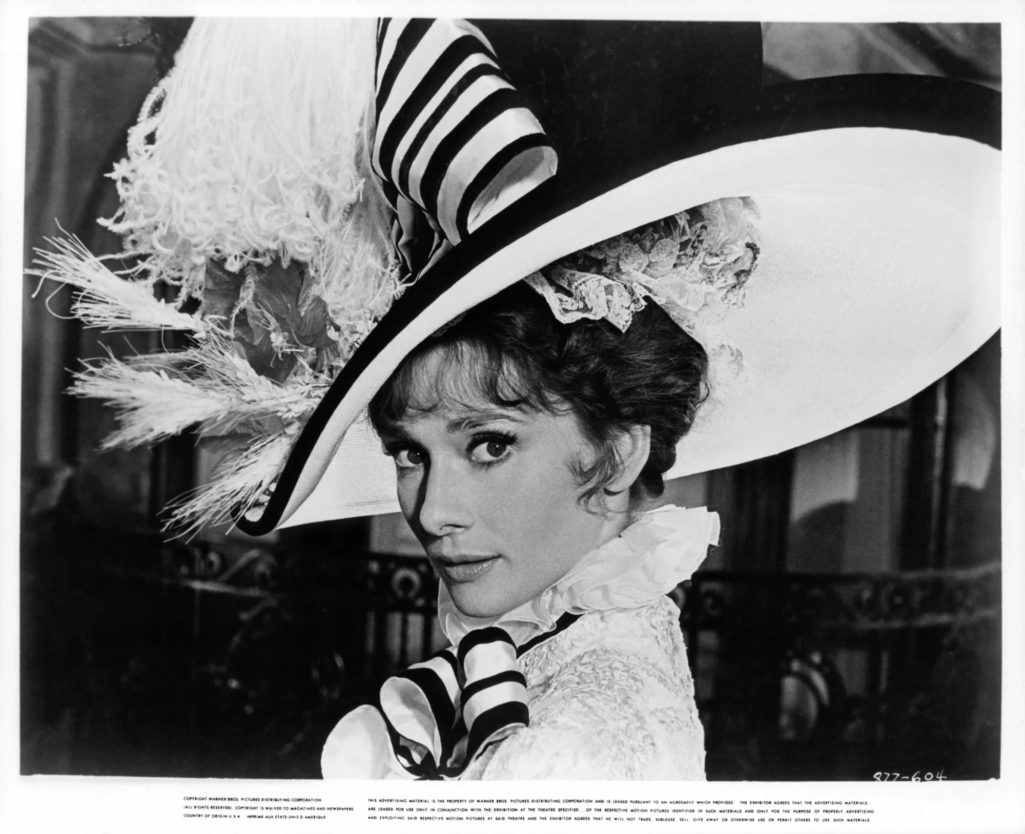 Audrey Hepburn, My Fair Lady filmindeki ikonik kostümüyle, büyük tüylü şapkası ve şık detaylarıyla dikkat çekici bir bakış atarken, beyaz perdenin unutulmaz anlarından birini yansıtan bu siyah beyaz fotoğrafta zarif ve etkileyici görünüyor.
