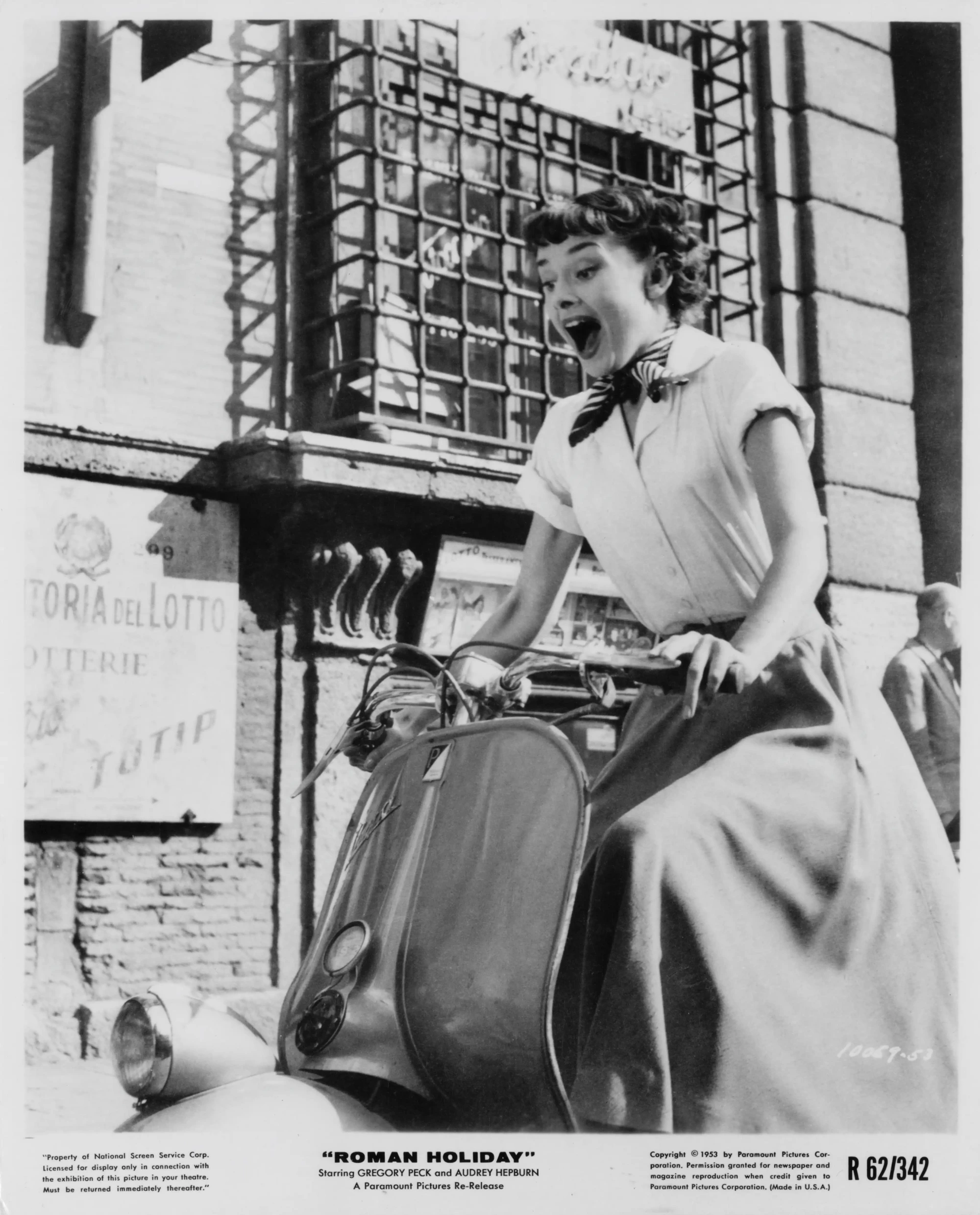Siyah beyaz bir fotoğrafta, Audrey Hepburn, Roman Holiday filminden prenses Ann karakteriyle, Roma sokaklarında bir scooter üzerinde şaşkınlıkla ağzı açık, boynunda fular ve açık renkli uzun eteğiyle özgürlüğün tadını çıkararak, unutulmaz anlardan birini yaşarken görülüyor.