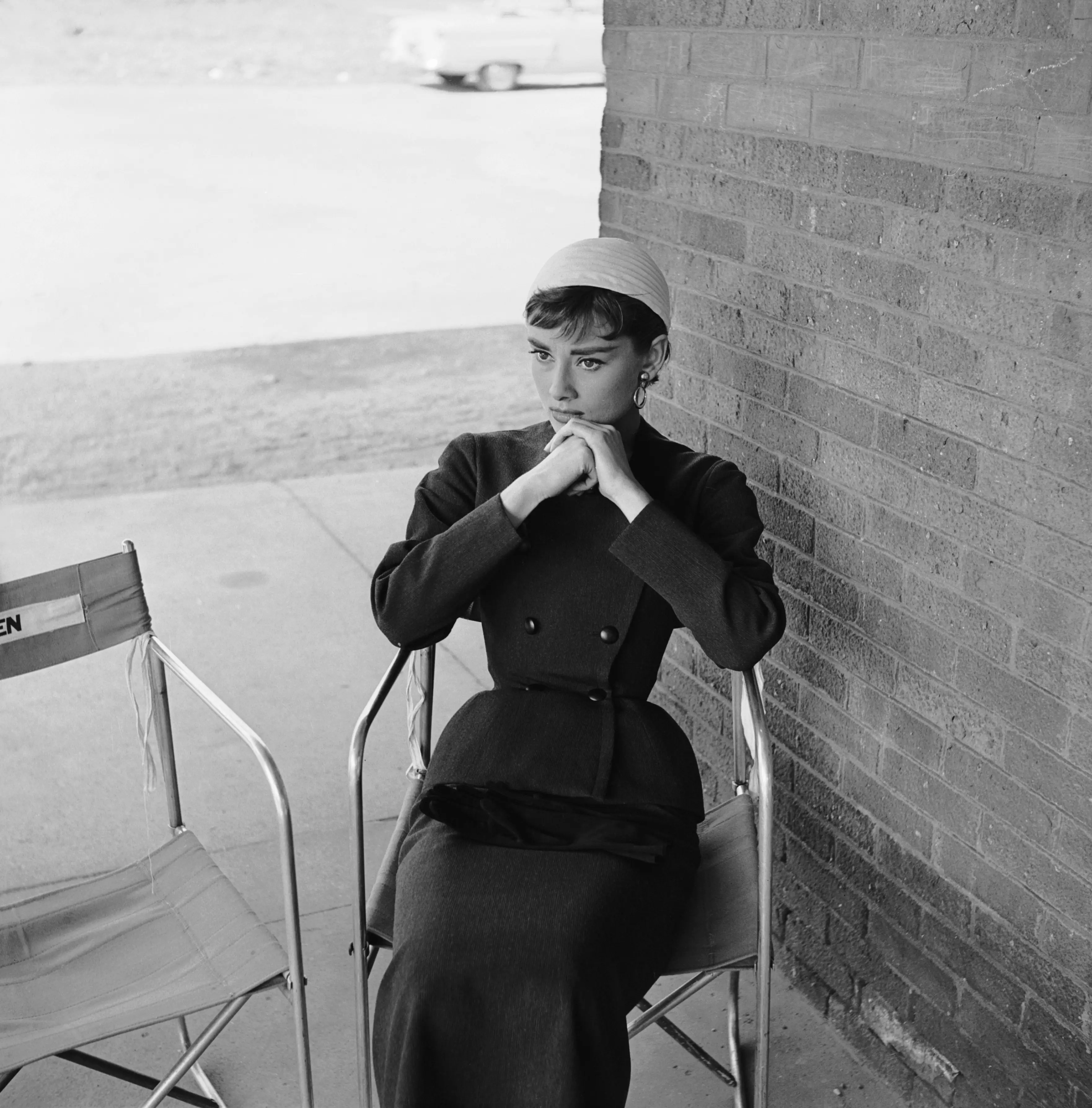 Sabrina filminin setinde, ikonik tarzıyla dikkat çeken Audrey Hepburn, koyu renk takım elbisesi ve açık renk şapkasıyla, ellerini çenesinin altında kavuşturmuş, düşünceli bir şekilde oturarak unutulmaz anlarından birini yansıtıyor.