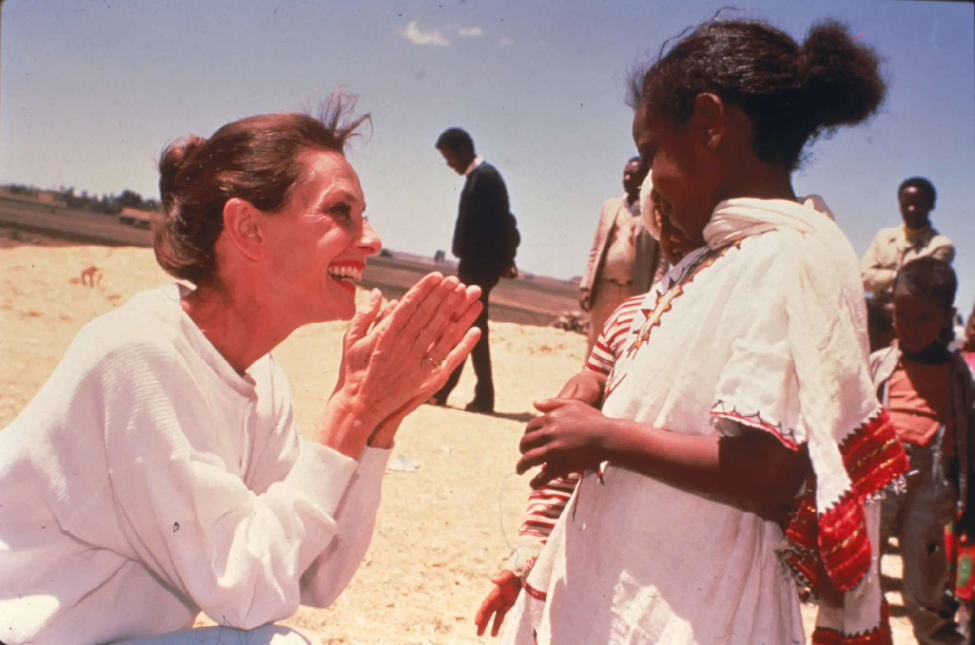 Audrey Hepburn, beyaz yakalı sade bir bluz giymiş, parlak bir gülümsemeyle eğilmiş, ellerini içtenlikle birleştirerek karşısındaki geleneksel kıyafetler içindeki Afrikalı çocuğun gözlerine bakarken, UNICEF elçiliği yaptığı yıllardaki bu unutulmaz anlardan birinde görülüyor.