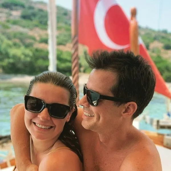 Güneş gözlüklü, mutlu yüz ifadeleriyle birbirlerine sarılmış Alp Kırşan ve eşi Zeynep Kırşan, bir teknenin güvertesinde, arka planda dalgalanan Türk bayrağı ve yeşil kıyılar eşliğinde deniz tutkularını ve mavi yolculuk keyiflerini yansıtan samimi bir anı paylaşıyorlar.