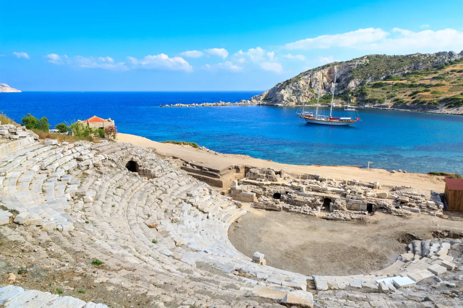Antik Knidos Tiyatrosu'nun kemerli kalıntıları ve basamaklı oturma yerlerinin önünde, masmavi denizin kucakladığı bir koyda zarif bir guletin salındığı bu büyüleyici manzara, Alp Kırşan'ın bahsedilen deniz tutkusunu ve mavi yolculuk rotalarının eşsiz güzelliğini gözler önüne seriyor.