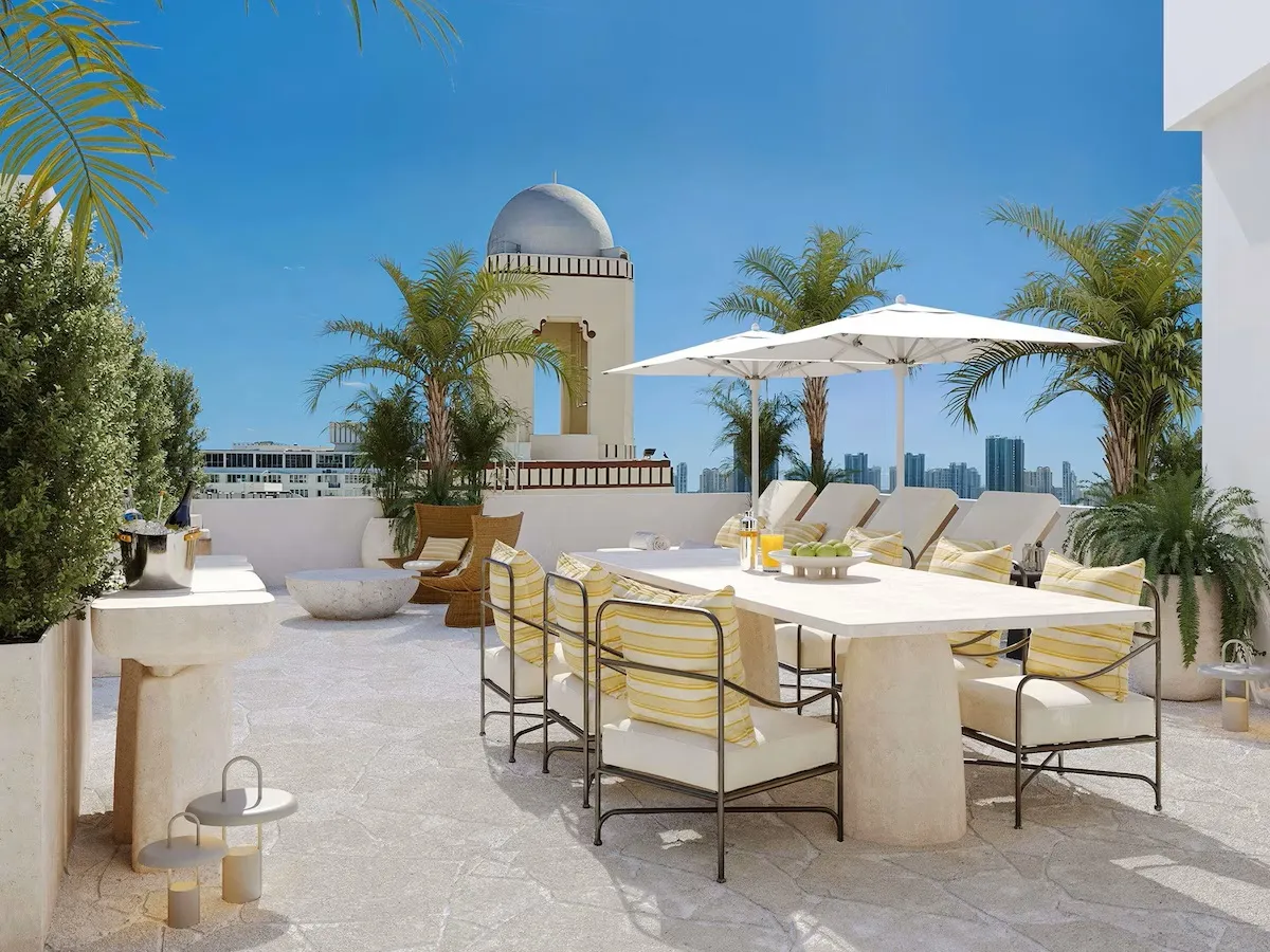 Miami South Beach'teki Delano Hotel'in güneşli lüks terasında, palmiye ağaçları ve uzaktaki şehir silüeti manzarası eşliğinde sarı çizgili minderli yemek masası ve şezlonglar keyifli anlar vadediyor.
