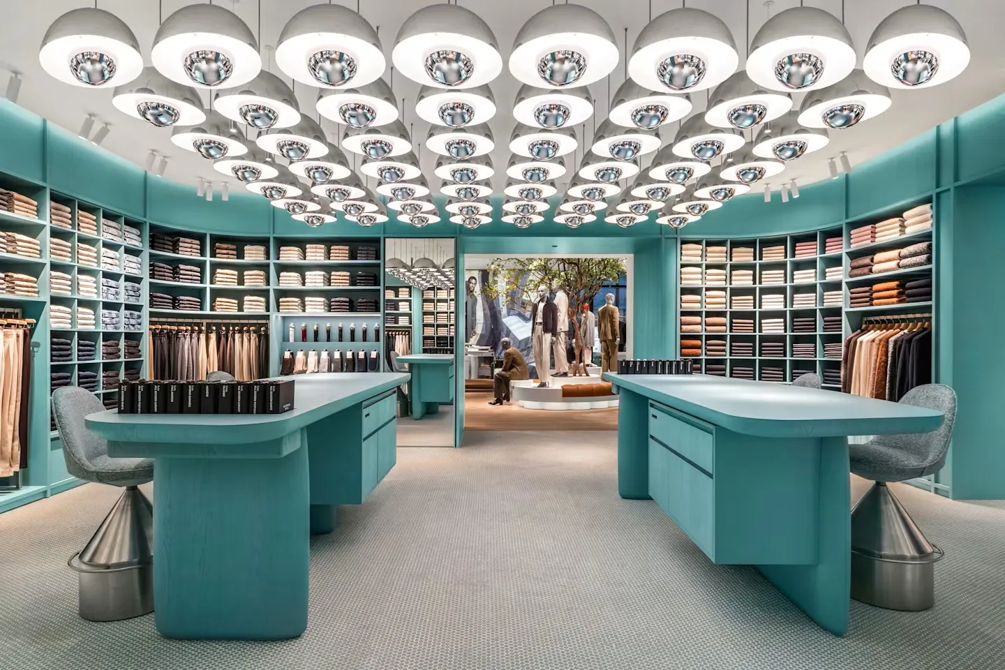 Miami Beach'te alışveriş için önerilen Suit Supply mağazasının modern iç tasarımı, turkuaz renkte duvarları, aynalı, kubbeli aydınlatma armatürleriyle dolu tavanı ve raflarda sergilenen özenle katlanmış gömlekler ile çeşitli takım elbiselerin kumaş örneklerini gösteriyor.