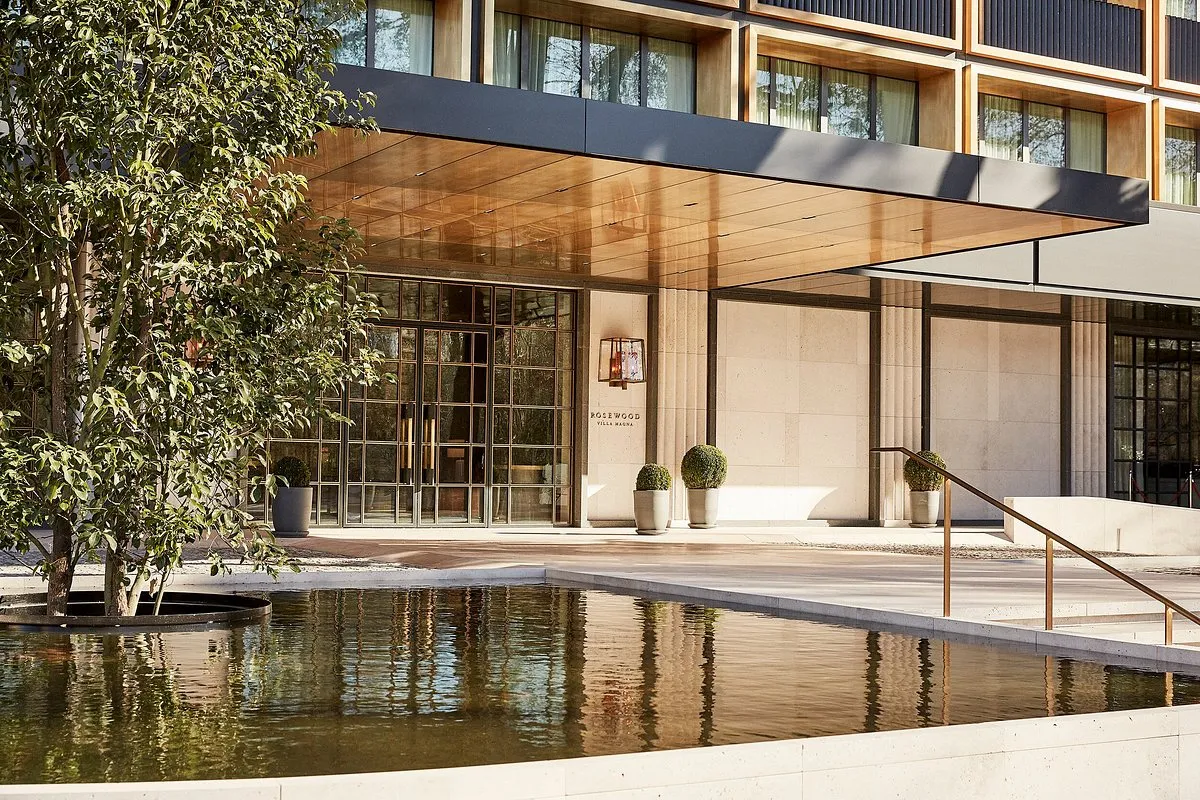 Madrid'in kalbinde yer alan Rosewood Villa Magna'nın modern ve zarif girişini gösteren bu geniş planda, ön planda yansımalı bir havuz, yemyeşil ağaçlar ve bronz renkli gölgelik altında parlayan şık cam kapılar otelin İspanya turizmine kapılarını açtığını vurguluyor.