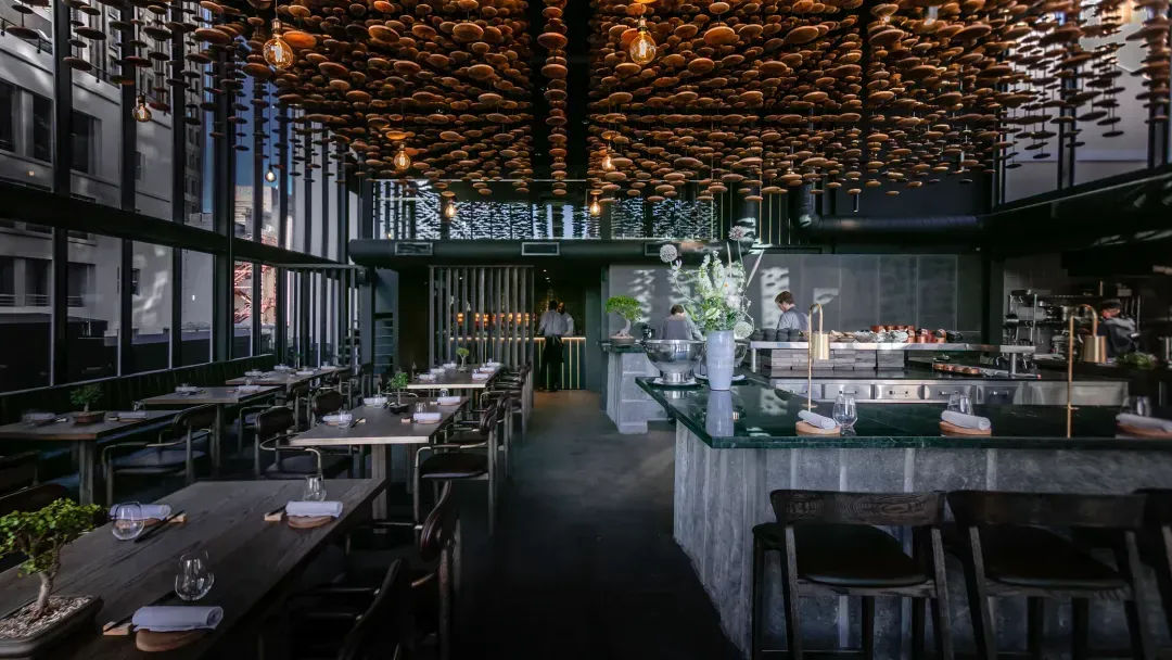 Tavanı yüzlerce küçük, kahverengi disk şeklinde süslemelerle bezenmiş, modern ve şık bir restoranın iç mekanı; sol taraftaki büyük cam pencerelerden şehir manzarası görünürken, sağda açık mutfak ve hazırlık alanı yer alıyor, bu tasarım Cape Town’daki gurme restoranların özgün ve sofistike atmosferini yansıtıyor.