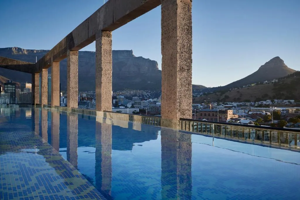 Cape Town'da konaklama deneyimini vurgulayan bu görüntüde, lüks bir otelin çatı katındaki sonsuzluk havuzundan, şehrin panoramik manzarası ve arka planda tüm ihtişamıyla yükselen Masa Dağı ile Lion's Head görülüyor.