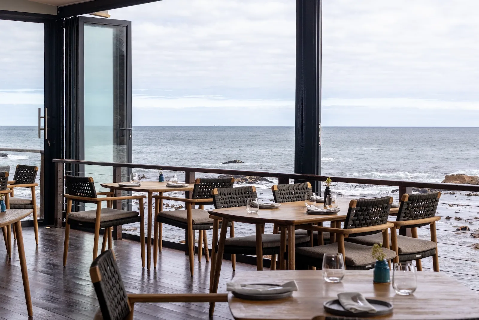 Cape Town'daki Tintswalo Atlantic otelinin okyanus manzaralı lüks restoranında, zarif ahşap masalar ve sandalyeler düzenlenmiş, cam kapılar ardında kayalık kıyıya vuran dalgalar ve geniş deniz ufku net bir şekilde görülüyor.
