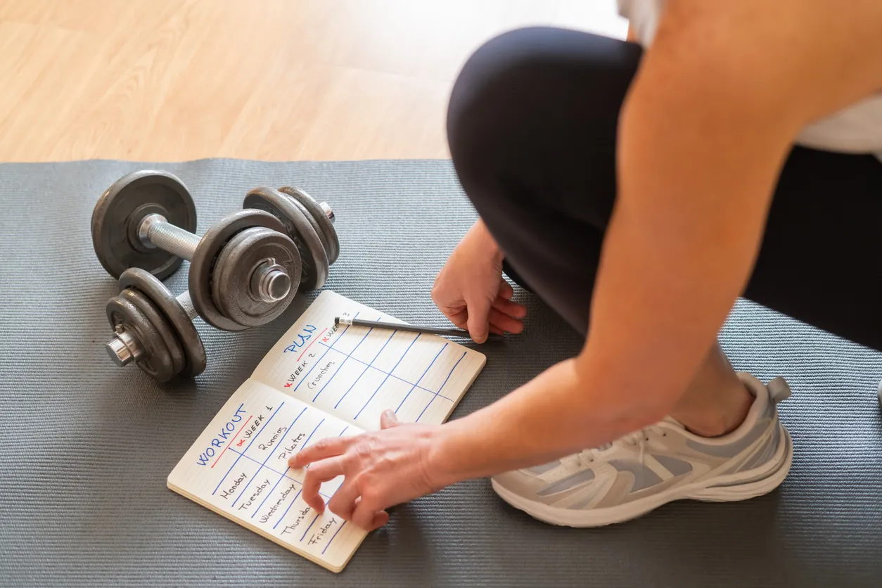 Evde spor programı planlamanın detaylarını gösteren bu görselde, spor matı üzerinde oturan bir kişi, önündeki açık defterine WORKOUT PLAN başlığı altında haftalık antrenmanlarını (koşu, pilates gibi) not alırken, yanında iki adet dambıl görülüyor.