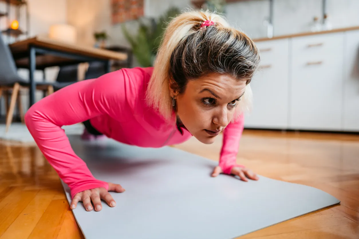 Parlak pembe uzun kollu spor tişörtü giymiş, saçları at kuyruğu yapılmış bir kadın, evde spor yaparken ahşap zemindeki açık gri yoga matı üzerinde şınav pozisyonunda, yüzünde kararlı bir ifadeyle odaklanmış durumda.