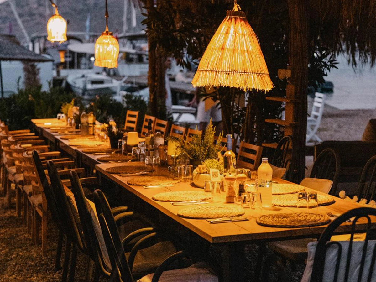 Kocabahçe Koyu'nda tekneyle ulaşılabilen bir restoran olan Kocabahçe Glamping'in, denize nazır akşam yemeği için sazdan büyük sarkıt lambaların sıcak ışığıyla aydınlatılmış uzun ahşap masasının ve etrafındaki ahşap sandalyelerin oluşturduğu davetkar ve doğal atmosferi.