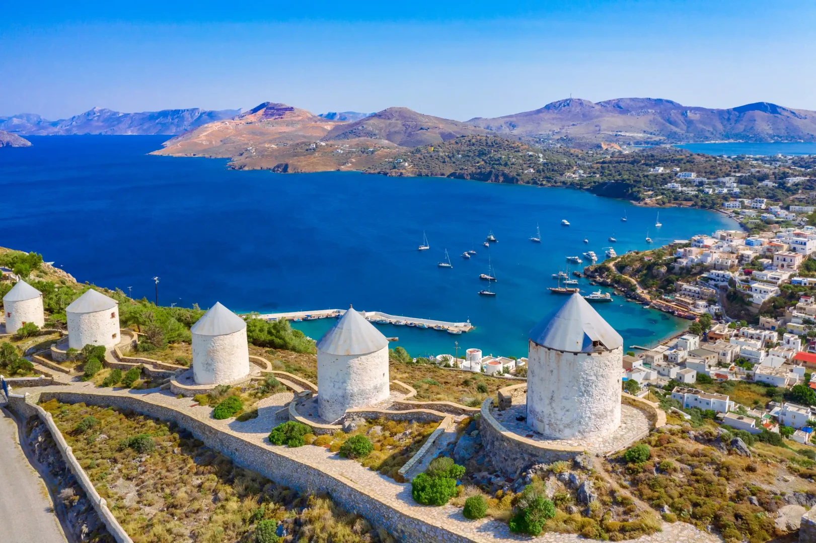 Leros Adası'nda, yamaç boyunca sıralanmış geleneksel beyaz yel değirmenlerinin önünden görünen, masmavi Ege Denizi'ne demirlemiş çok sayıda yelkenlinin ve kıyıya yayılmış beyaz evlerin oluşturduğu huzurlu bir mavi yolculuk manzarası.