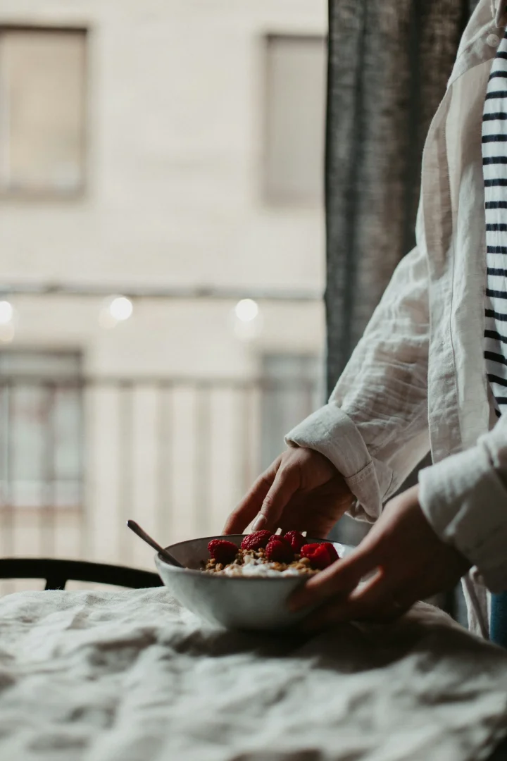 Açıklamalı bir mindful beslenme örneği olarak, keten örtülü bir masaya taze frambuaz ve granolayla süslenmiş yoğurt kasesini özenle yerleştiren bir kişinin elleri, yemeğe başlamadan önce yavaşlamanın ve duyuları devreye sokmanın önemini vurguluyor.