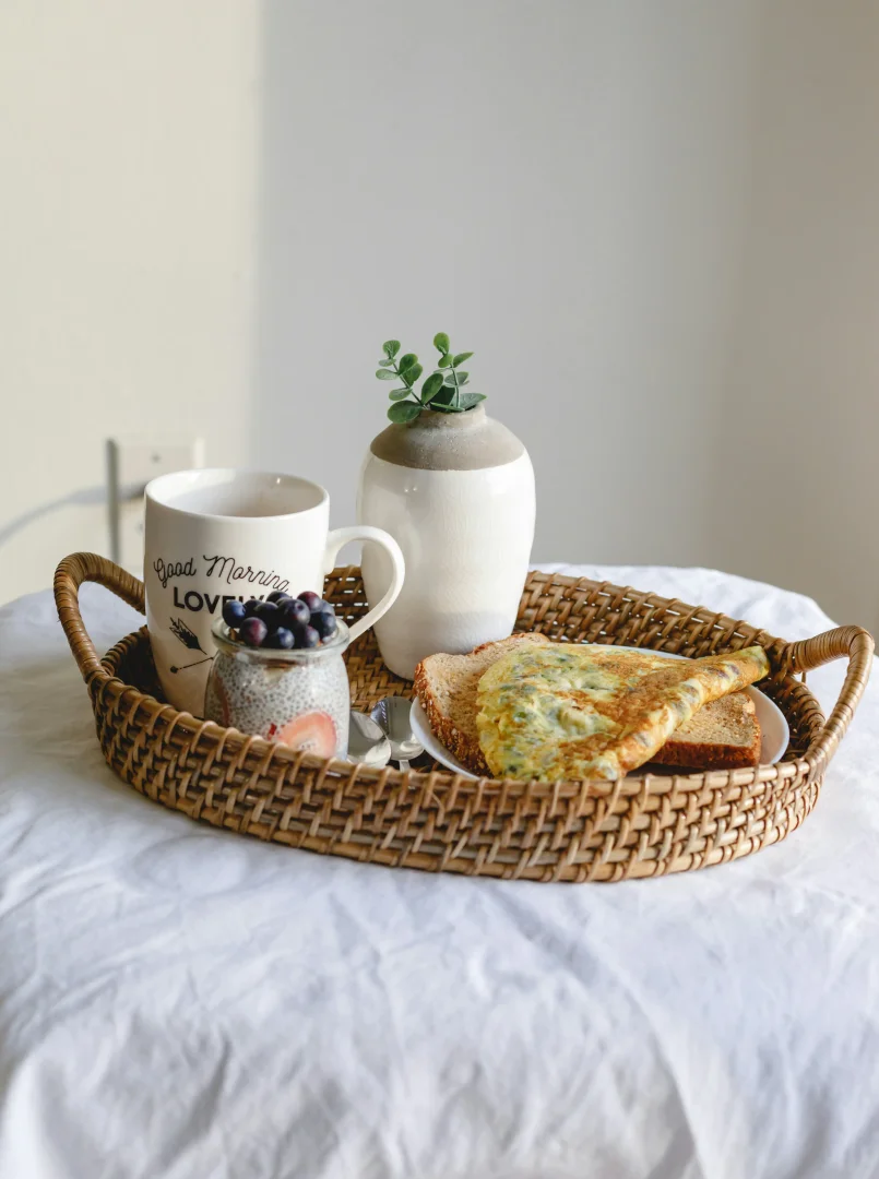 Hasır bir tepside, üzerinde Good Morning Love yazılı beyaz bir kupa, yaban mersini ve çilekli chia puding, yeşillikli omlet ve iki dilim kızarmış ekmek gibi çeşitli yiyeceklerin düzenli ve davetkar bir şekilde sunulduğu bir kahvaltı, mindful beslenme pratiklerinin dinginliğini ve yavaşlama adımlarını temsil ediyor.