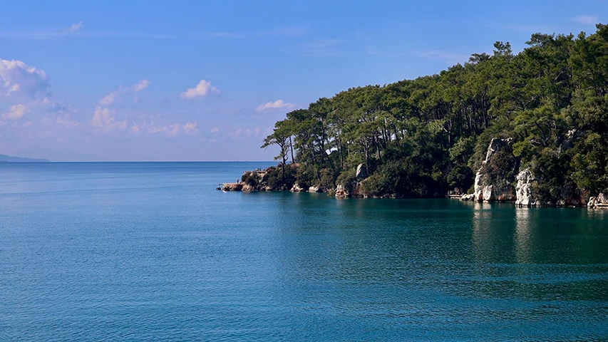 Muğla'nın Akyaka bölgesinde, çam ağaçlarıyla kaplı yemyeşil yamaçların berrak turkuazdan laciverte uzanan denize karıştığı kayalık kıyı şeridi ile üzerinde hafif bulutların süzüldüğü açık mavi gökyüzünün oluşturduğu doğal güzellikte bir manzara.