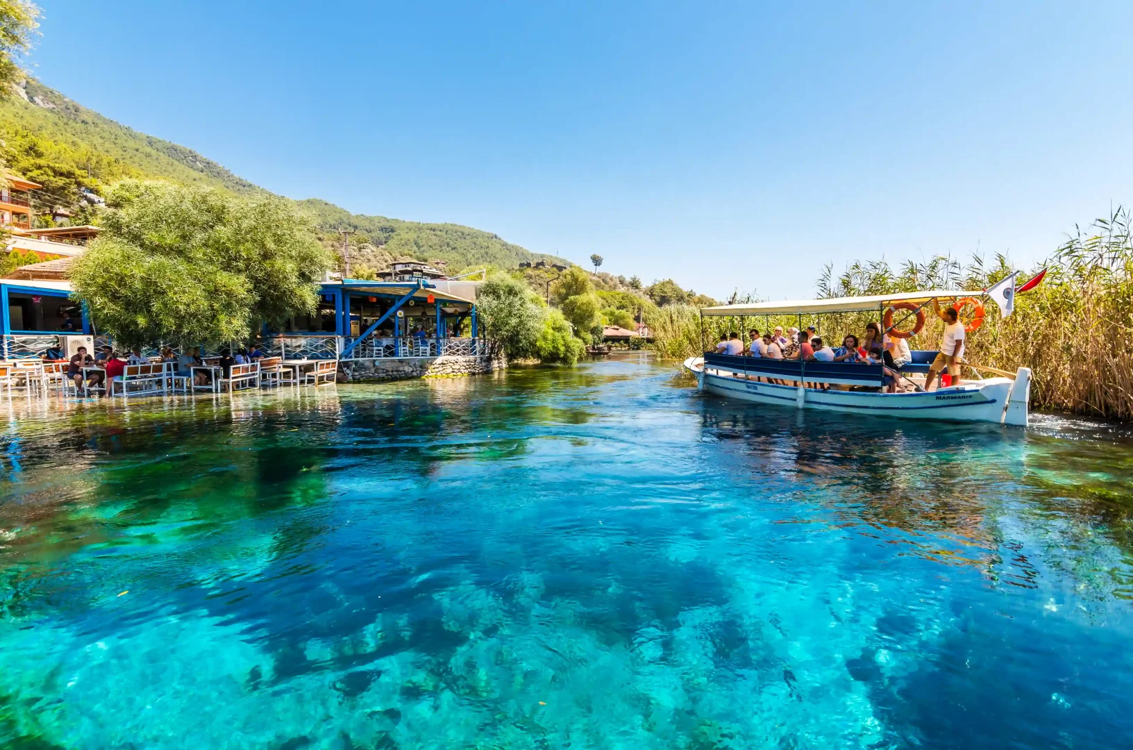 Akyaka'da, Gökova Körfezi road trip rotasının öne çıkan duraklarından Azmak Nehri'nin berrak turkuaz sularında ilerleyen tekne, yemyeşil doğa ve nehir kenarındaki restoranlarla çevrili huzurlu bir atmosferde yolculara unutulmaz anlar yaşatıyor.